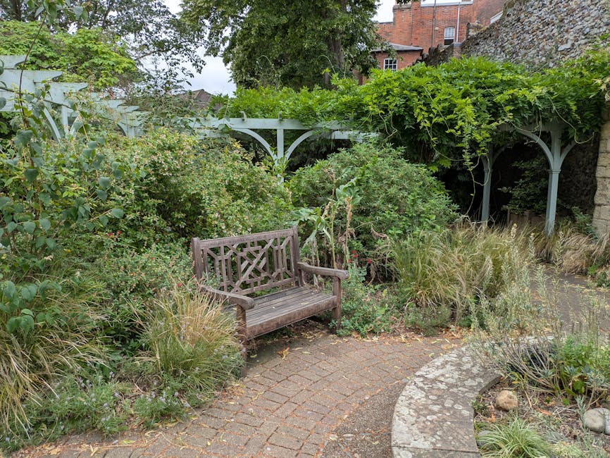 A small outdoor garden area featuring a wooden bench placed on a curved brick pathway. Surrounding the bench are various lush green plants, including low-growing grasses with wispy seed heads, broad-l
